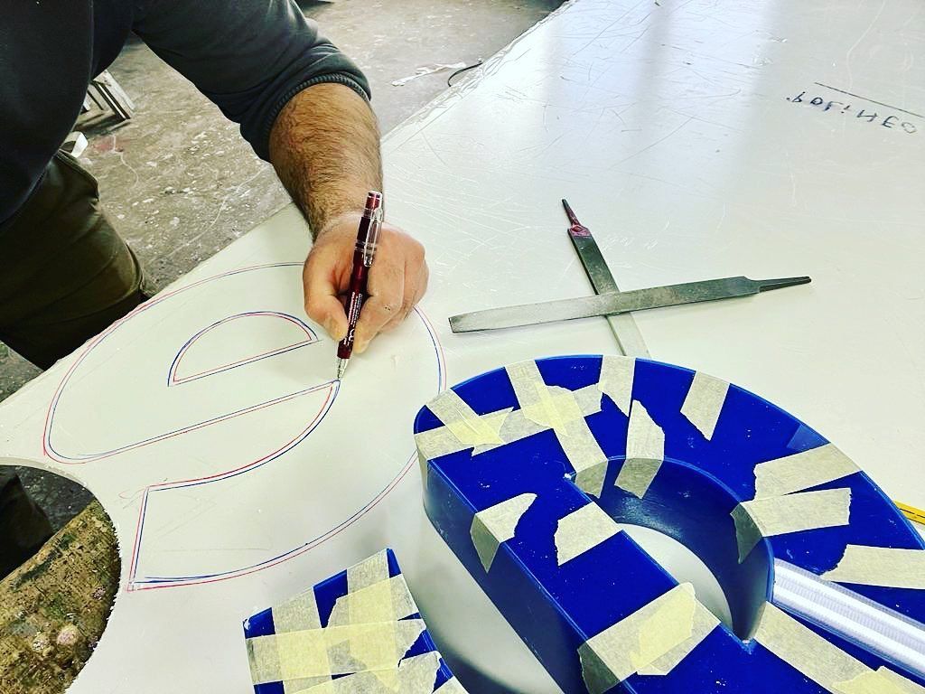 Persona che traccia il contorno di una lettera su una superficie bianca, con accanto una lettera blu ricoperta di nastro adesivo.