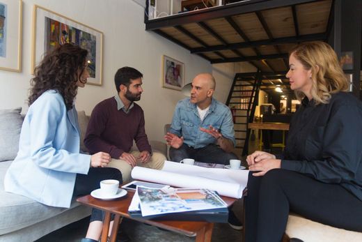 People sitting at a wooden table, one writing in a notebook. Sunlight streams in.