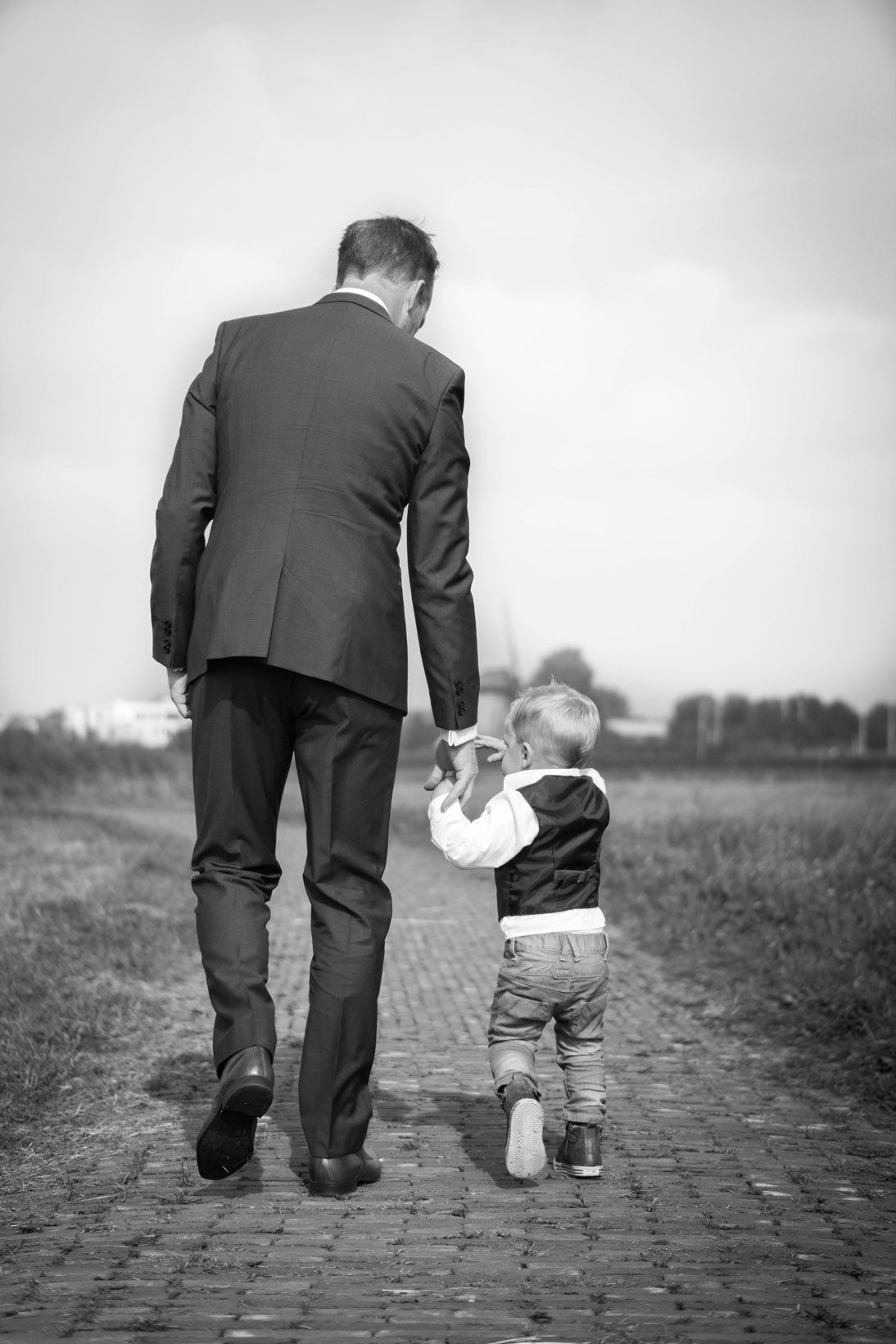 Man in suit holds hands with a young child walking on a brick path. Black and white.