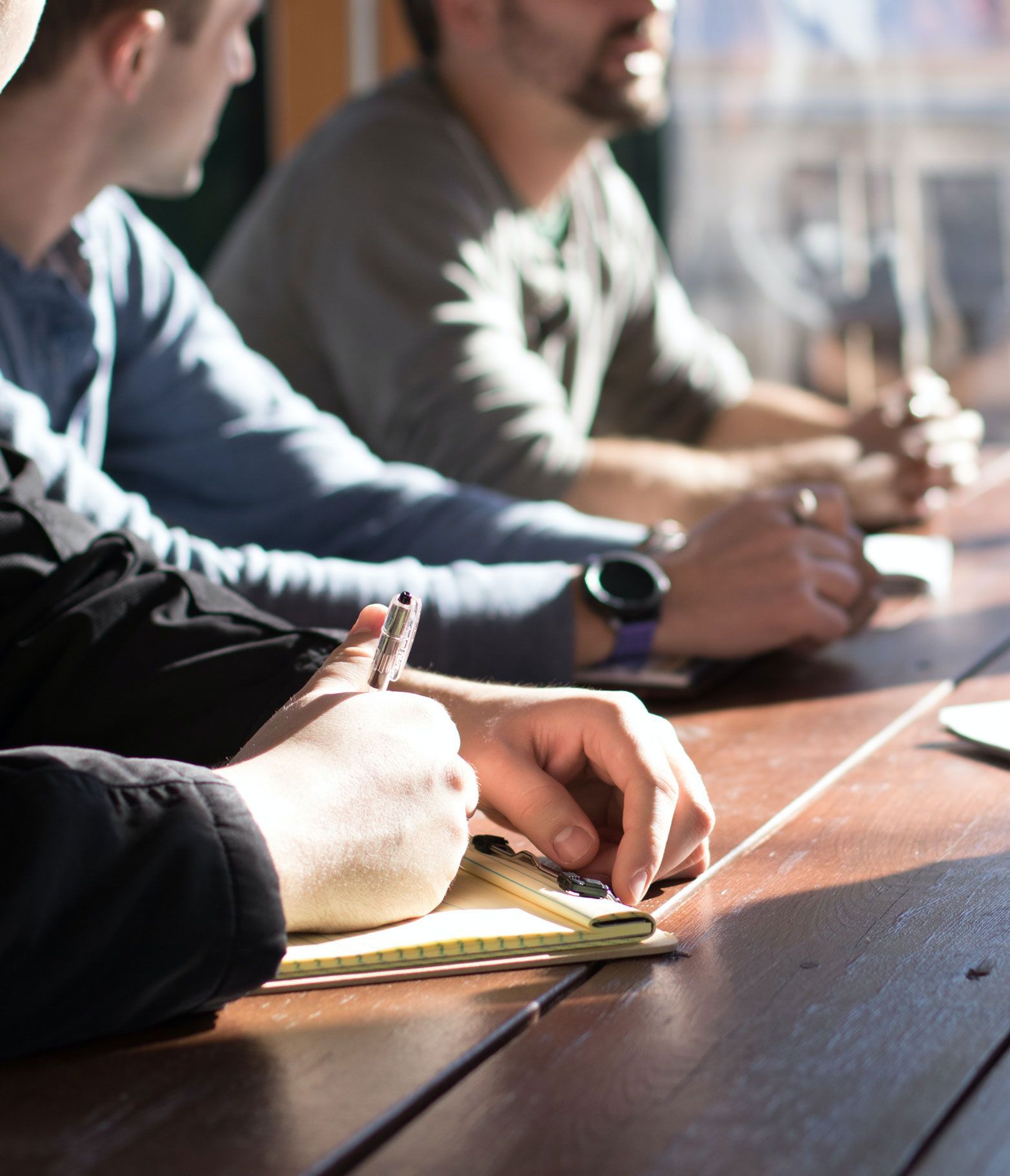 People sitting at a wooden table, one writing in a notebook. Sunlight streams in.