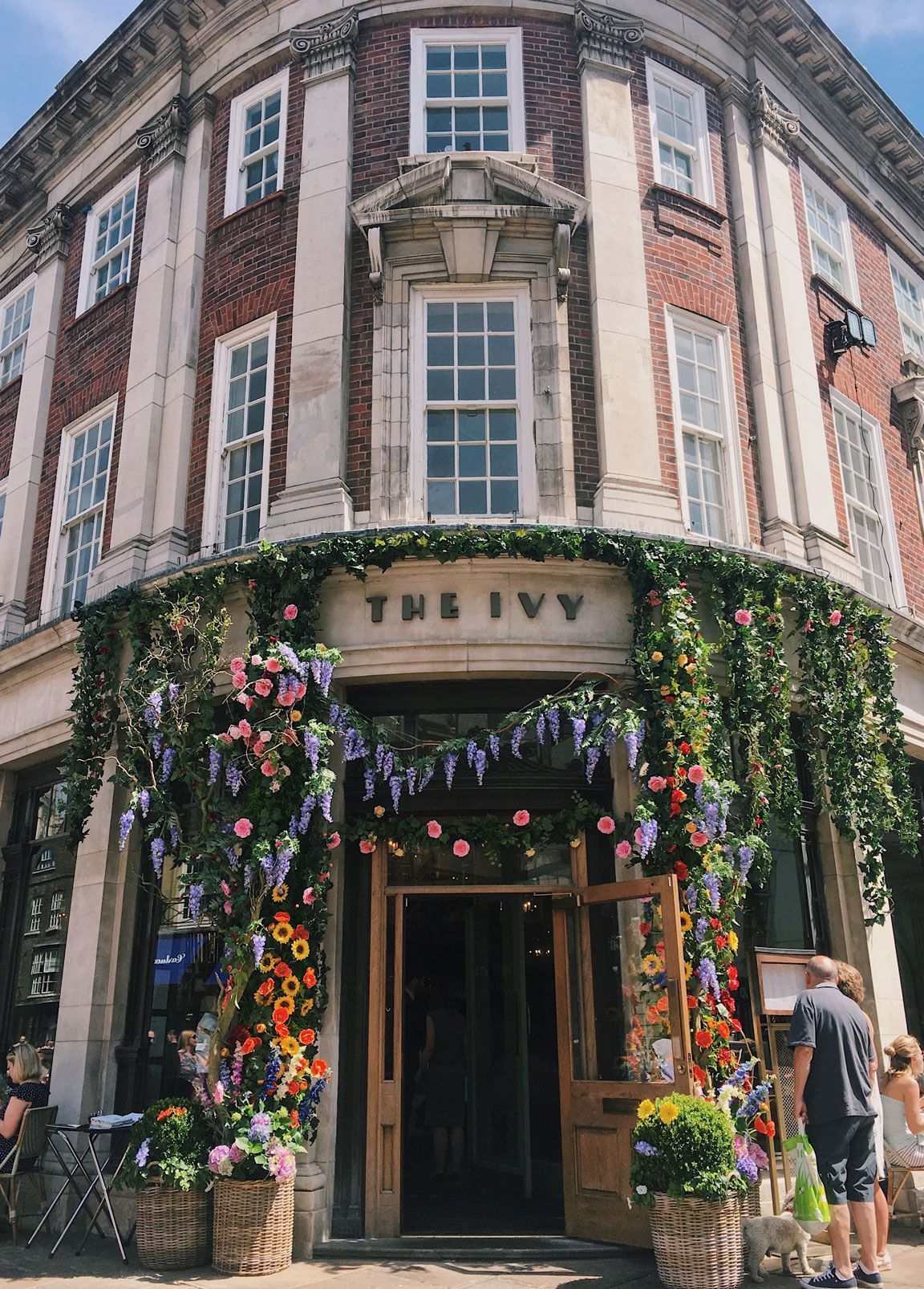 The Ivy restaurant entrance, decorated with flowers. Red brick building with large windows. Person looking at menu.