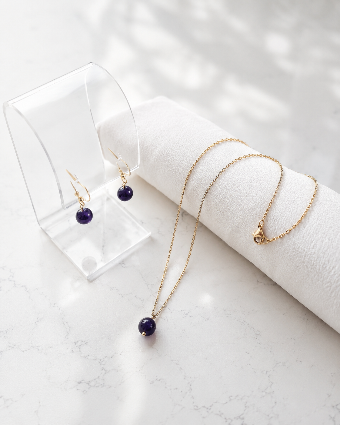Goudkleurige RVS ketting en oorbellen met paarse amethist kralen op een lichte marmeren achtergrond.