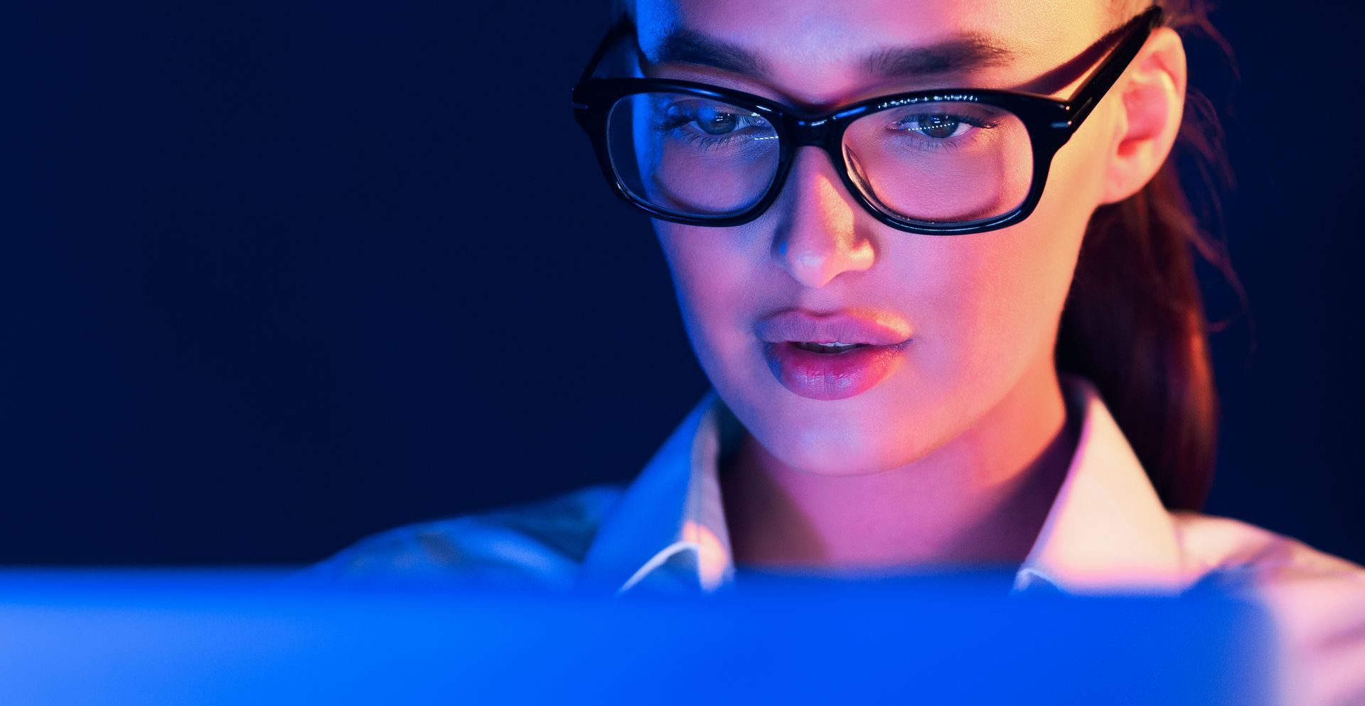 Woman with glasses looking intently at a screen, illuminated by blue and purple light.