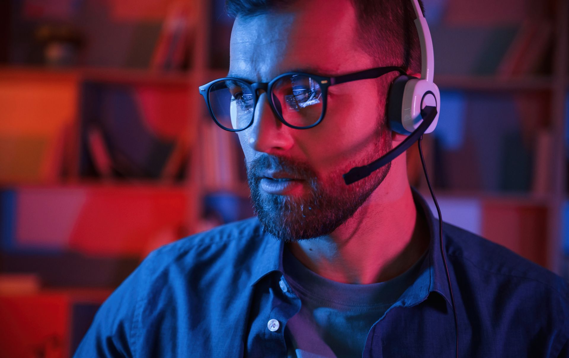 Man with glasses and headset in a room lit with red and blue light, looking at a screen.