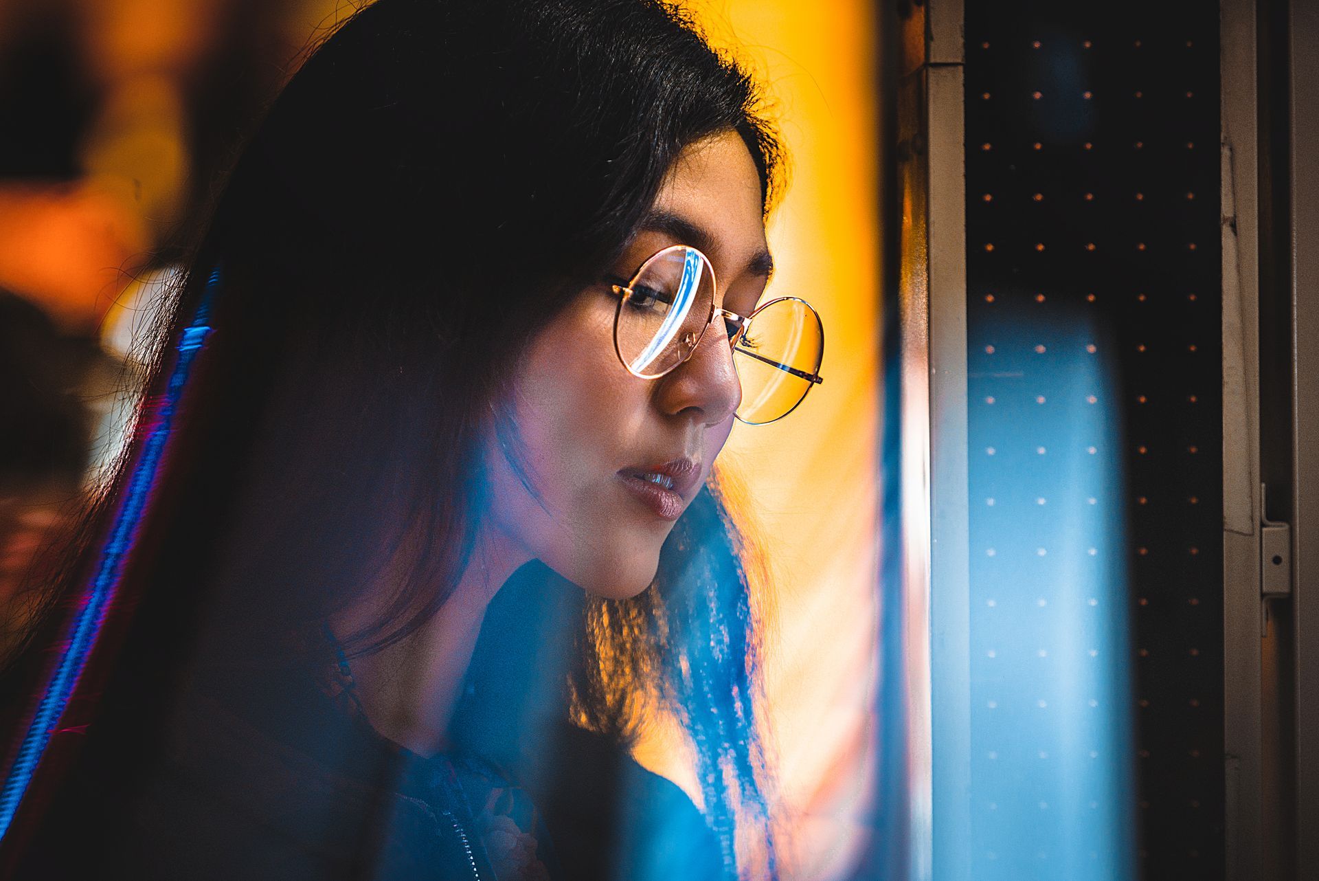 Woman with glasses looking at a brightly lit display, surrounded by blue and orange light.