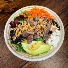 A bowl of salad with meat , avocado , carrots and other vegetables on a wooden table.