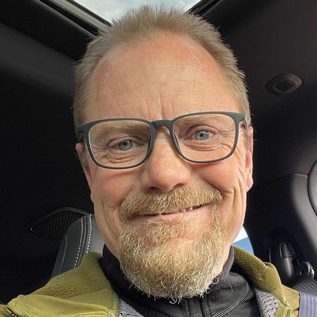 A man with a beard wearing sunglasses and a black shirt is smiling.