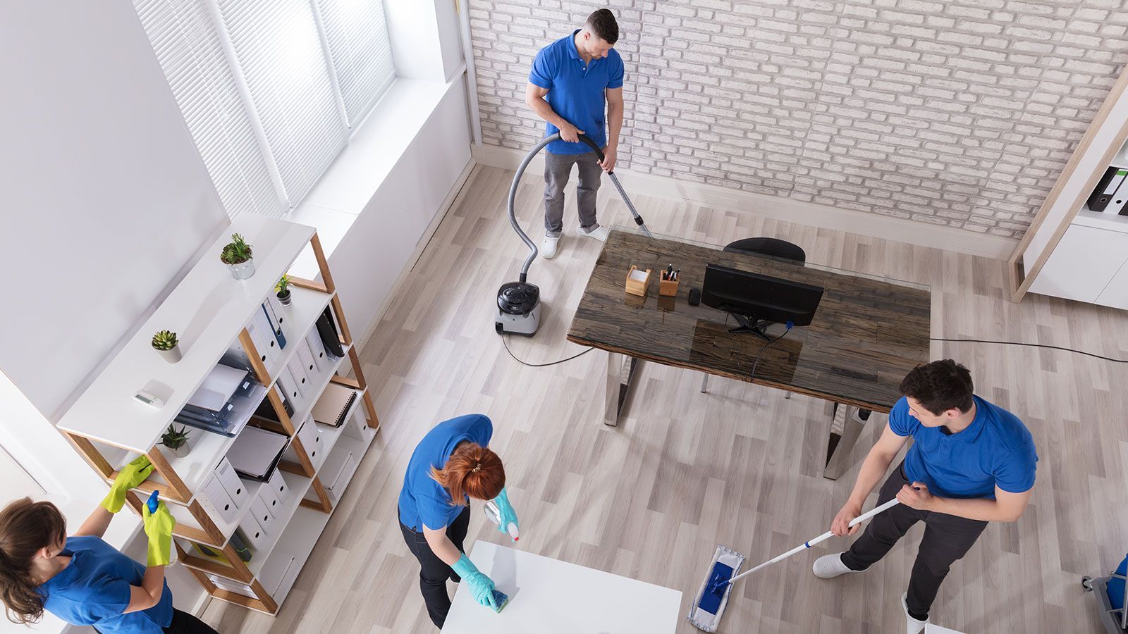 Office cleaning crew in blue shirts work: one vacuums, two mop, and two clean surfaces.