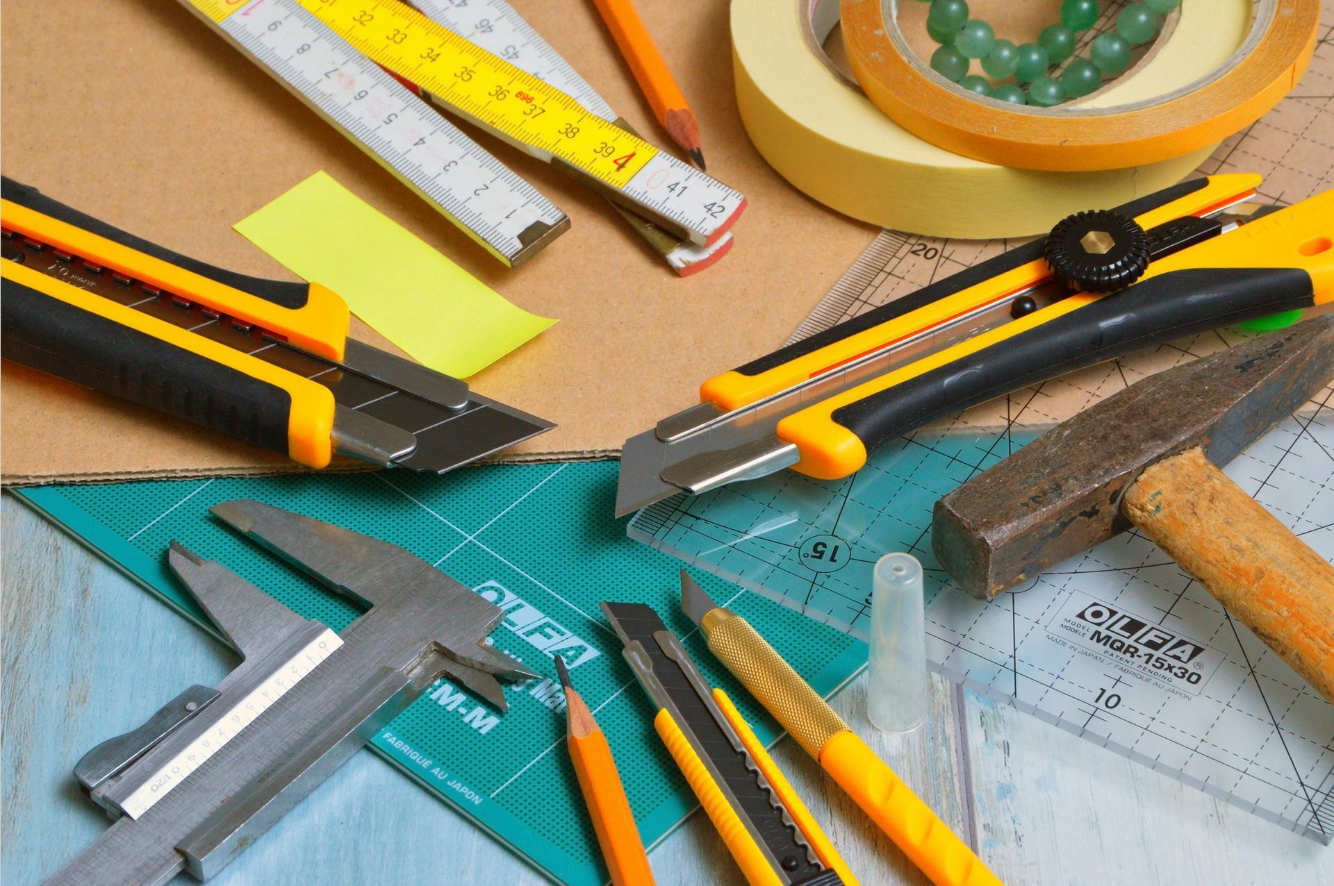 Close-up of craft tools on a table, including utility knives, a ruler, pencil, tape, and a hammer, suggesting a workshop or crafting setup.