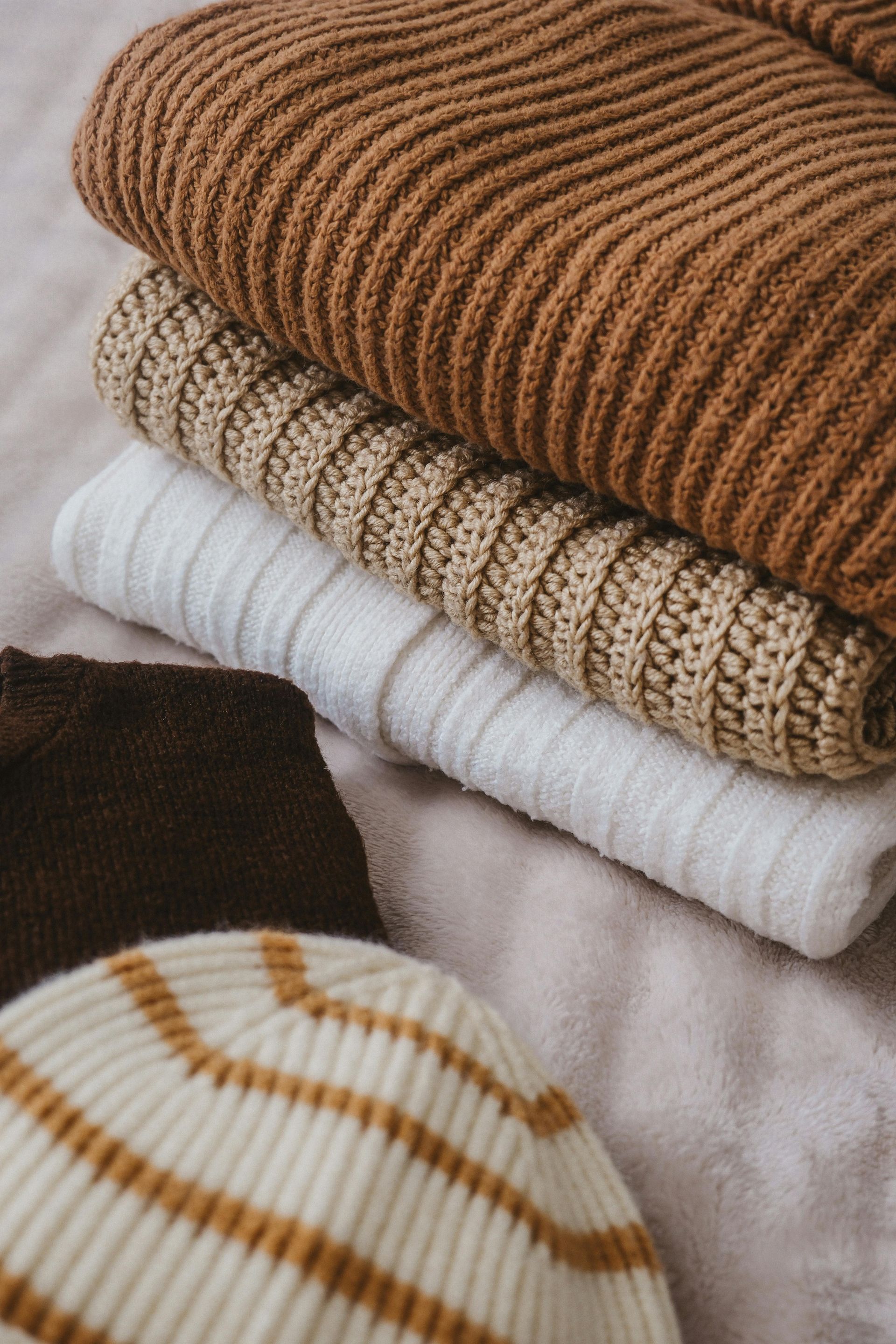 Stack of folded sweaters in shades of brown, cream, and white; a striped hat is at the bottom.