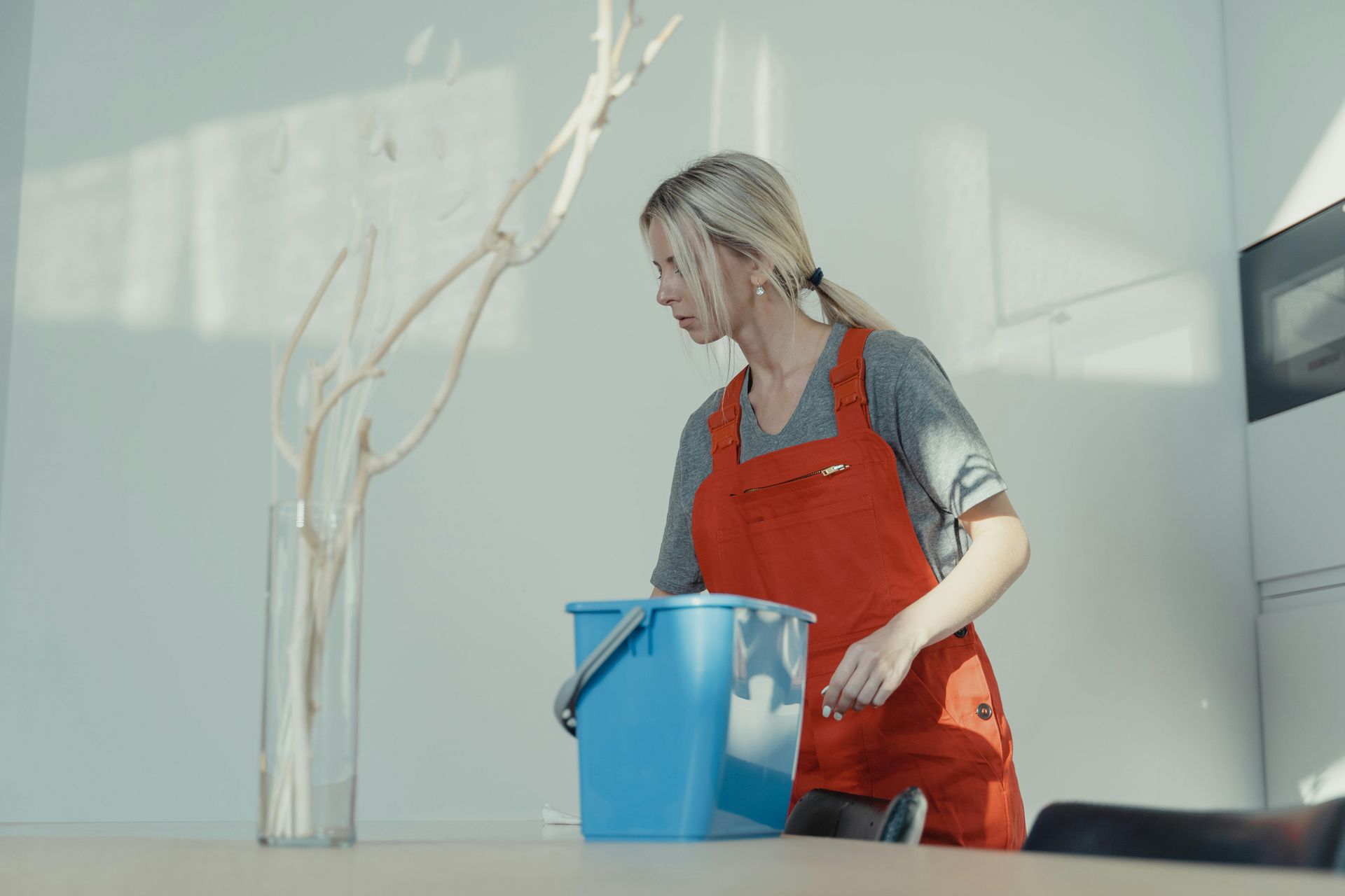Blonde woman in red apron cleans a table in a white room, holding a blue bucket near a tall vase with branches.