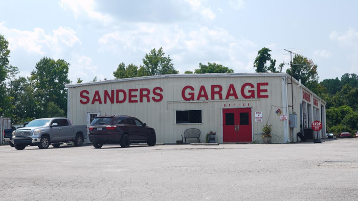 Sanders Garage building with signage, red soda machine, and flatbed tow truck on a sunny day.