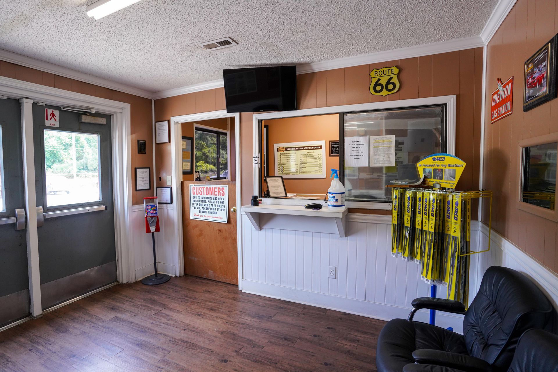 An interior view of a lobby with wood-paneled walls, a service window, a rack of yellow car parts, and a black armchair.