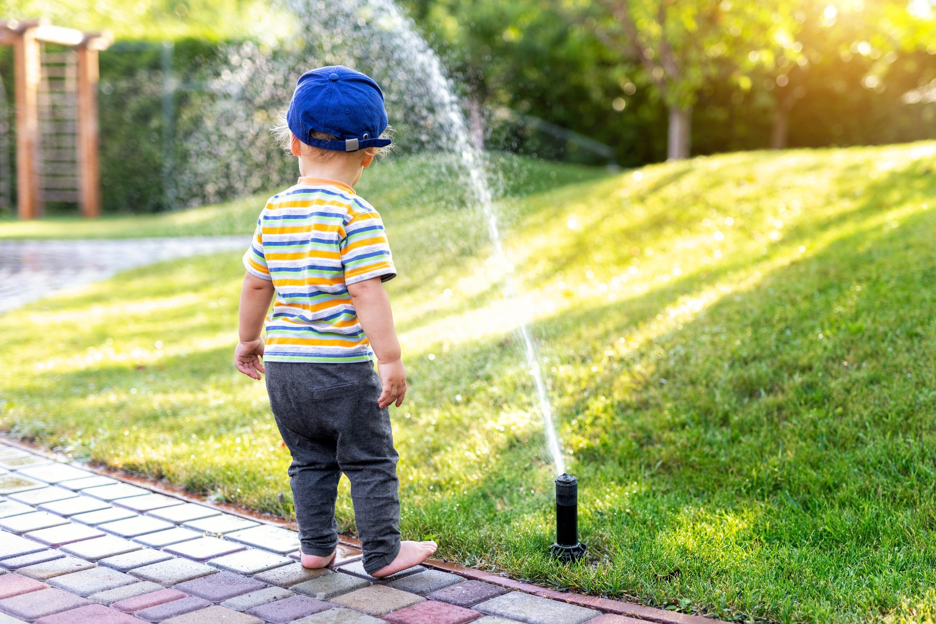 Child standing on a brick path watching a sprinkler spray water across a sunny lawn