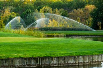 Green lawn with water sprinklers spraying arcs near a pond and trees in the background Green lawn with water sprinklers spraying arcs near a pond and trees in the background