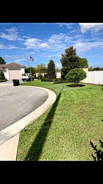 Suburban street with green lawn, curved sidewalk, white fence, trees, and a blue sky with clouds.