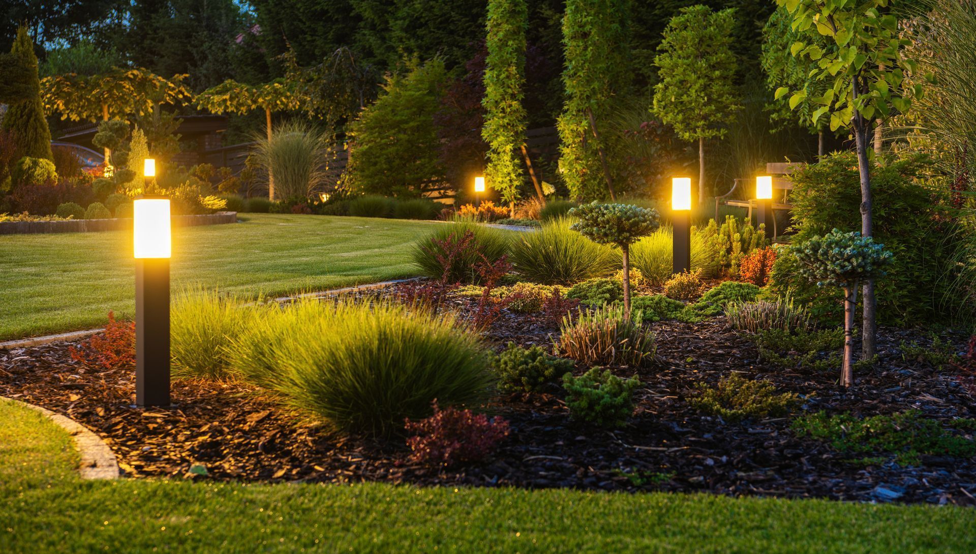 Lit garden path with glowing lanterns, shrubs, and trees at dusk
