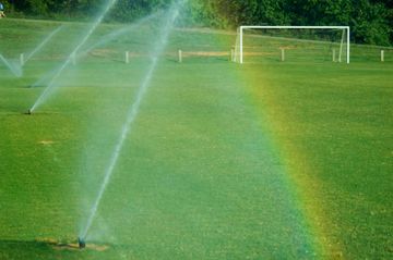 Sprinklers watering a green soccer field with a goal in the background, creating a rainbow.
