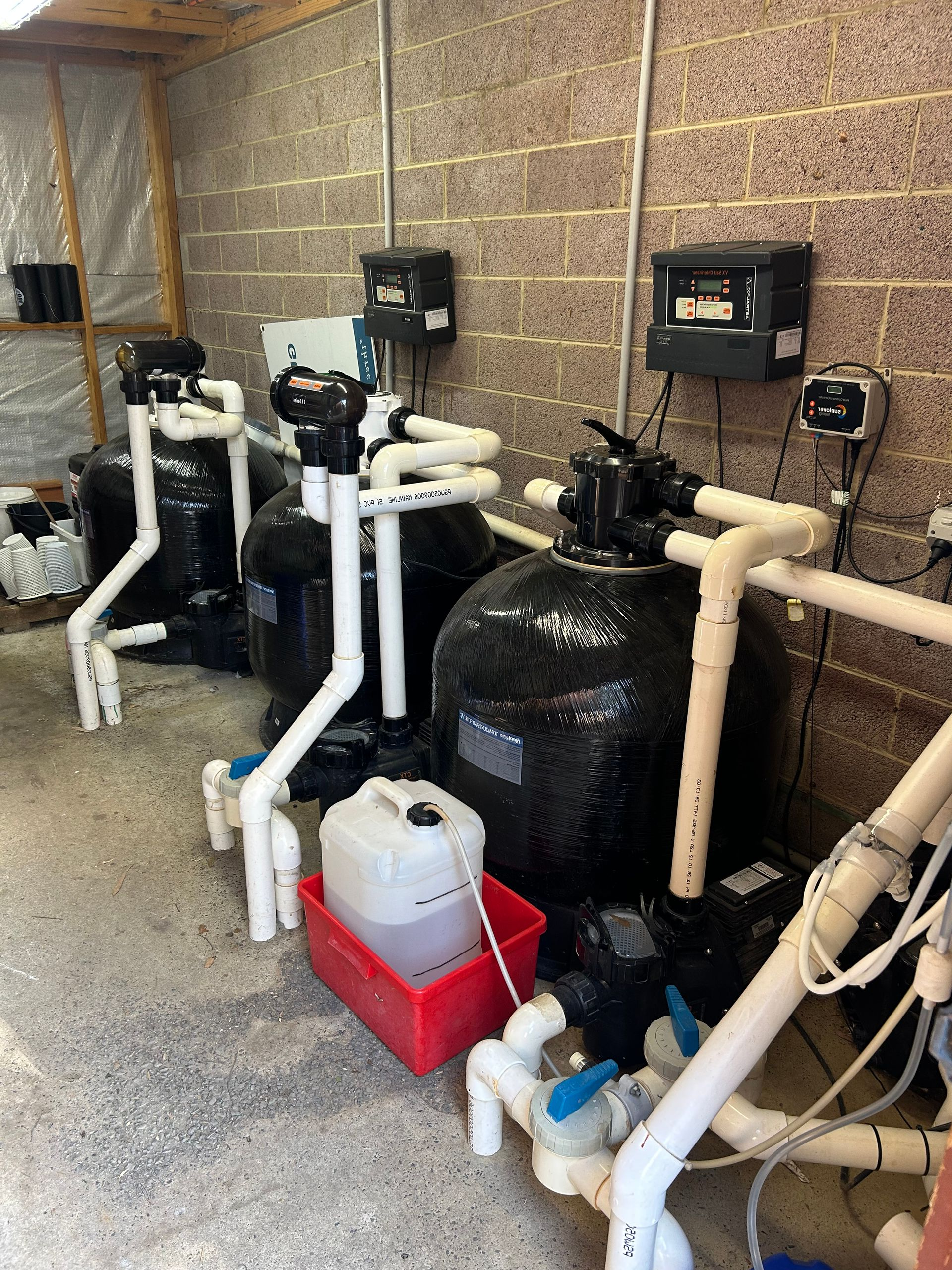 Row of pool pumps and filters in a utility room with beige block walls and concrete floor