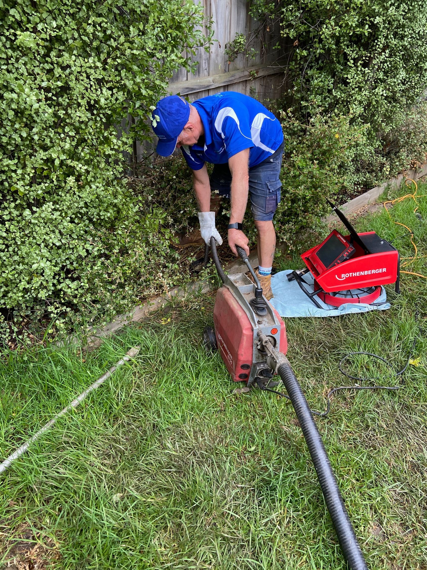 Technician in blue uniform repairing a hedge trimmer beside a hedge and red tool case outdoors