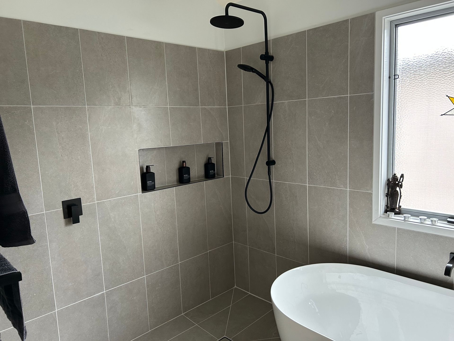 Modern bathroom shower with gray tiled walls, black fixtures, and a white bathtub beside a frosted window