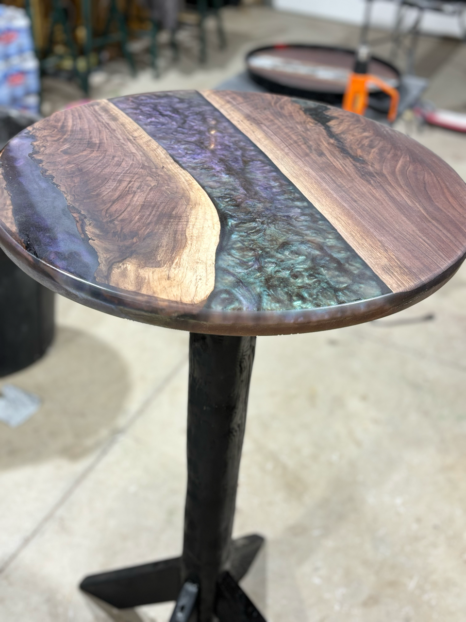 A round table with a live edge wooden top and a custom epoxy inlay and a black base is sitting on a concrete floor.