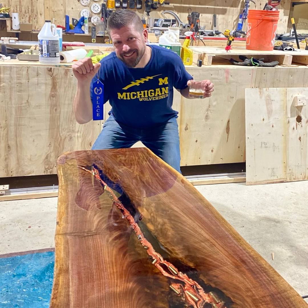 Jordan Timmerman, owner of Timmberman's Woodworking, in a michigan shirt is standing next to a wooden table displaying a first place ribbon