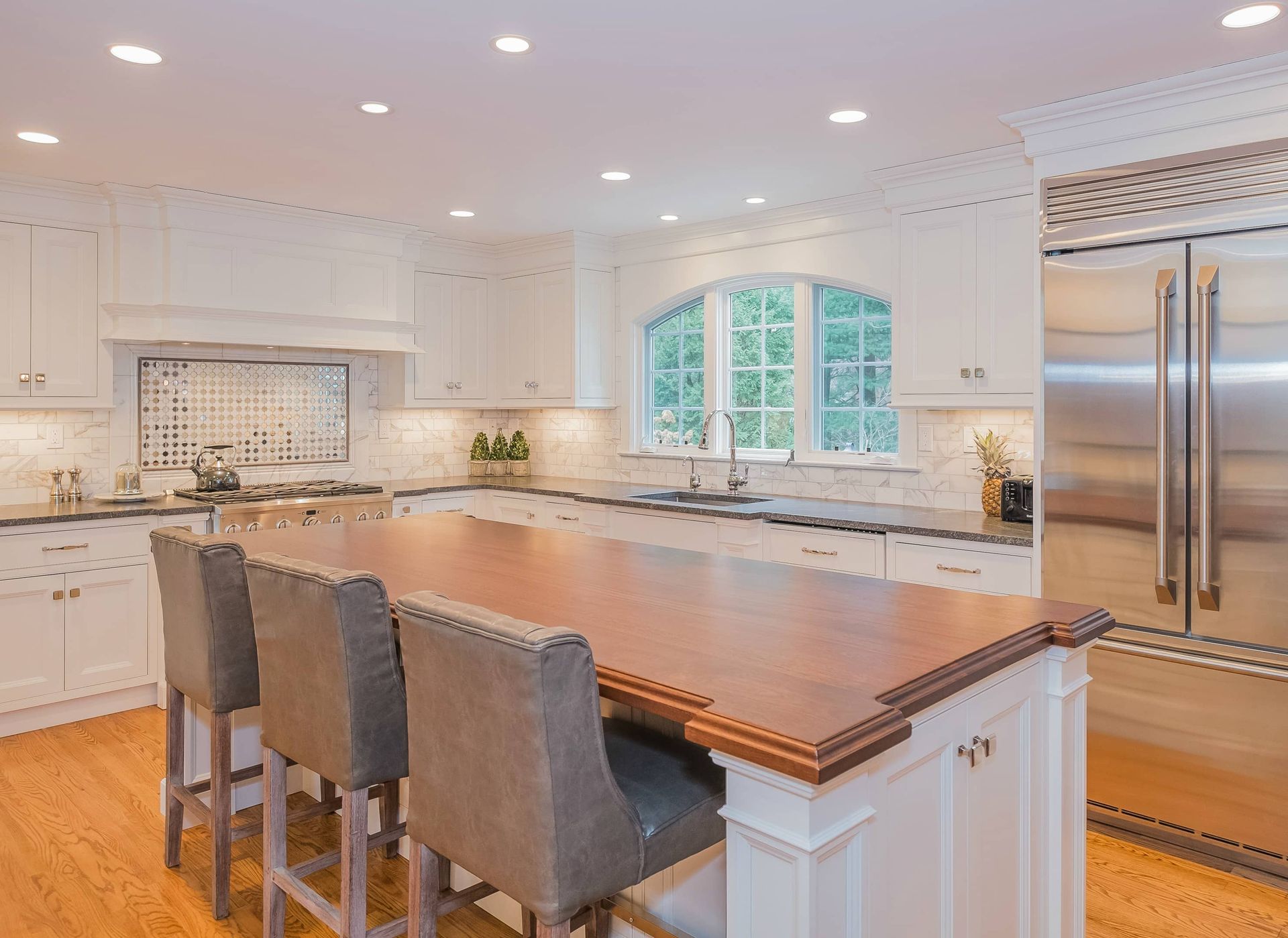 A bright, white kitchen featuring a large wooden-topped island with three gray upholstered bar stools.