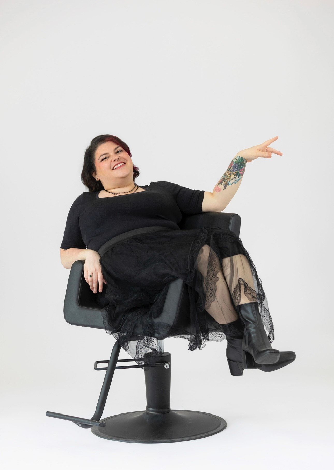 Woman in black outfit and boots, seated in salon chair, gesturing with her hand.