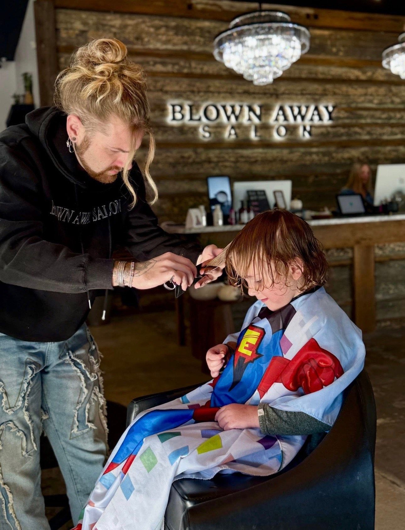 Barber cutting hair of a child wearing a superhero cape in a salon.