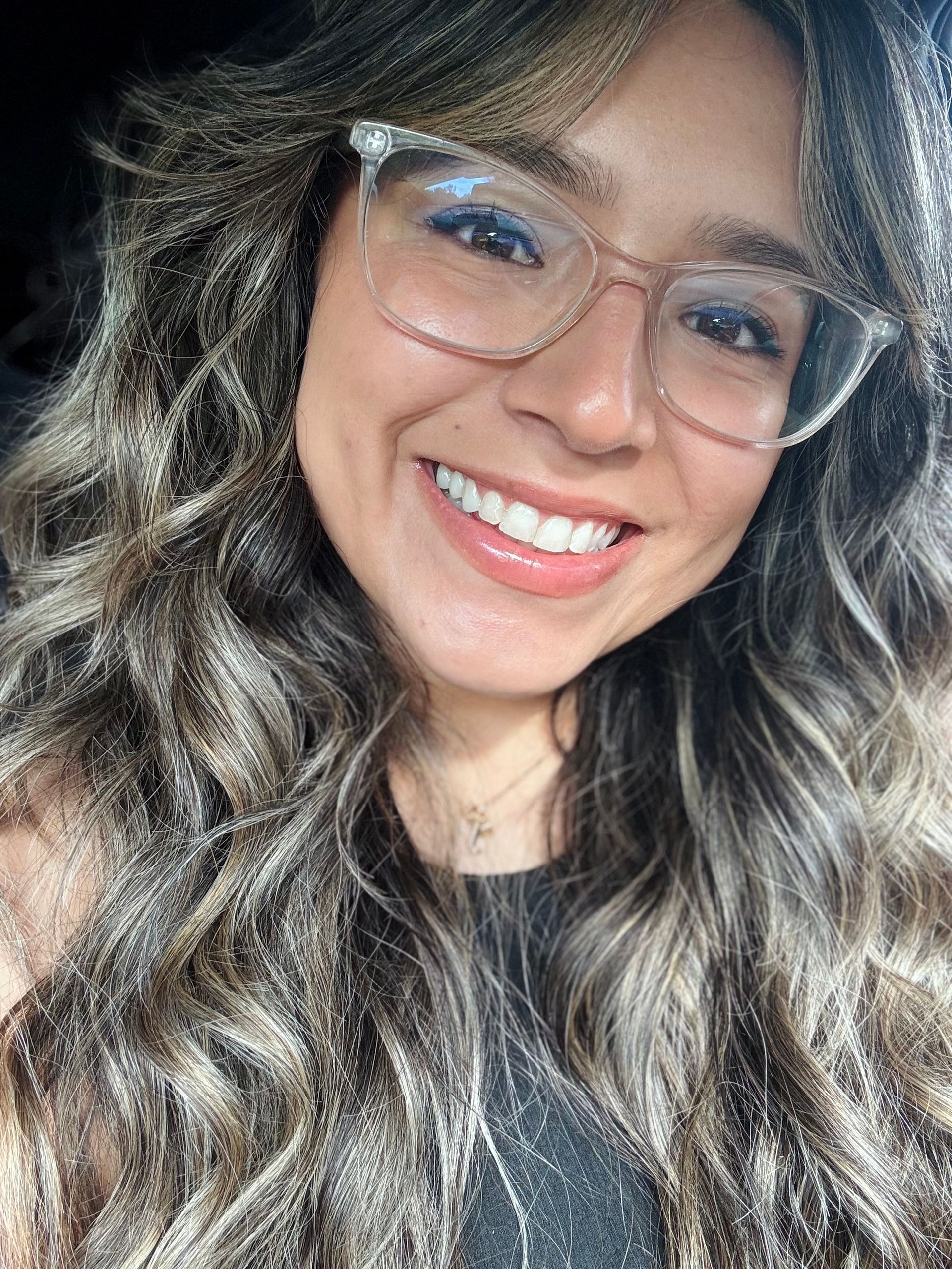 Woman smiling, wearing glasses. Long, wavy, highlighted brown hair.