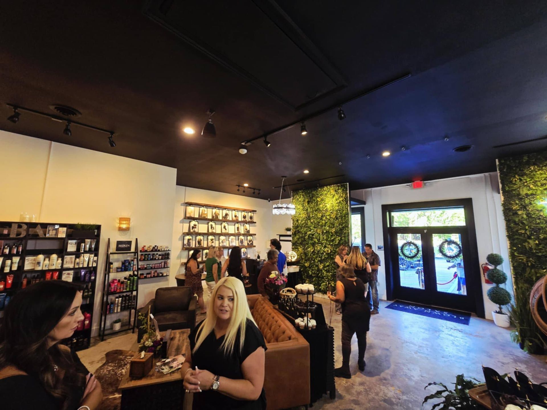 Interior of a shop with customers, products on shelves, and a faux-green wall. Black ceiling, people talking.