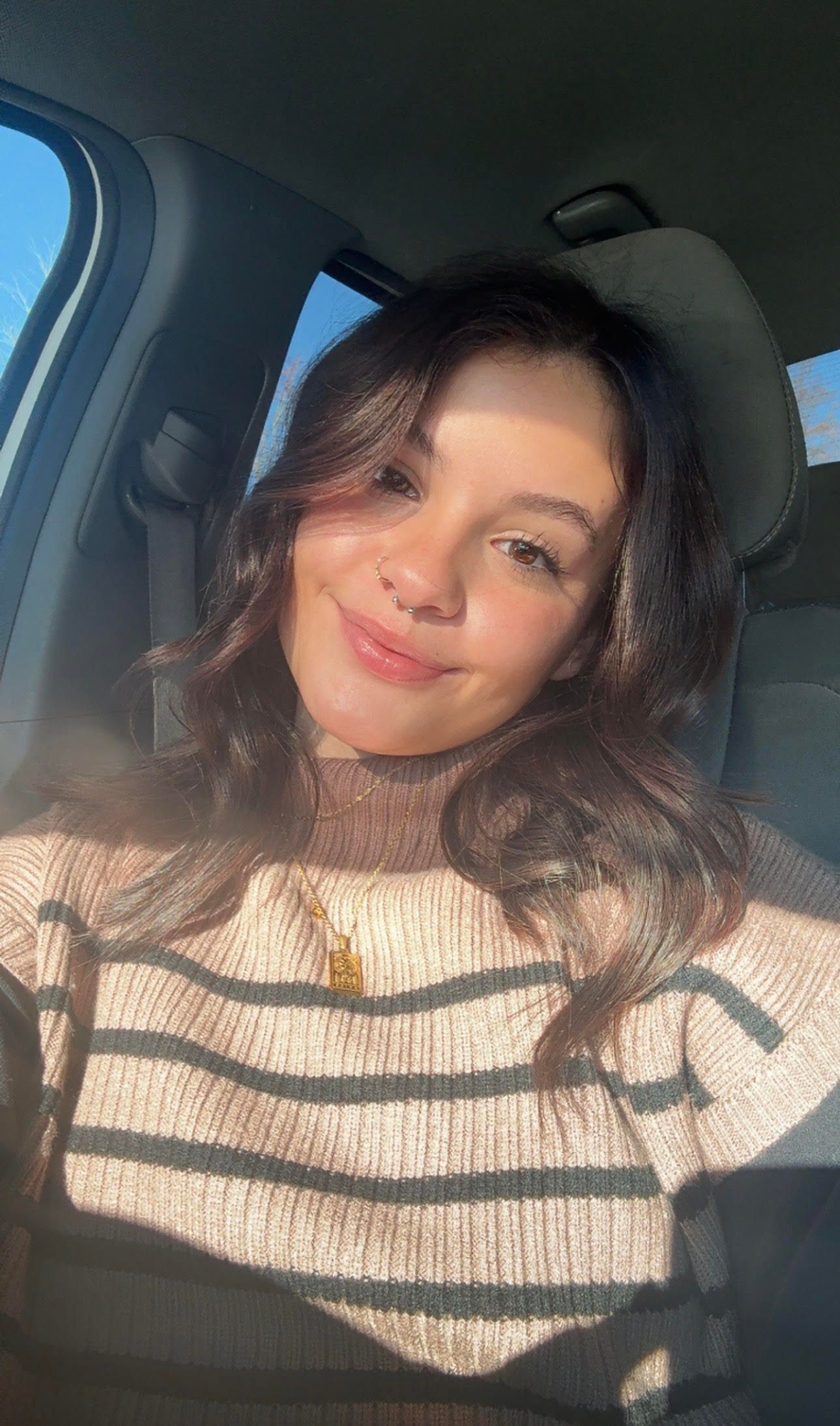 Woman in a car smiles, facing the camera. Wearing a striped sweater, sunlight brightens her face.