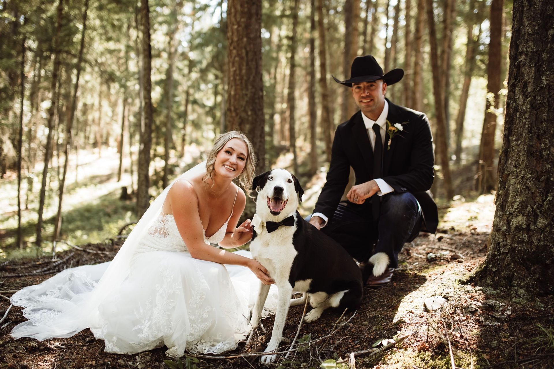 A bride and groom are posing for a picture with their dog in the woods.