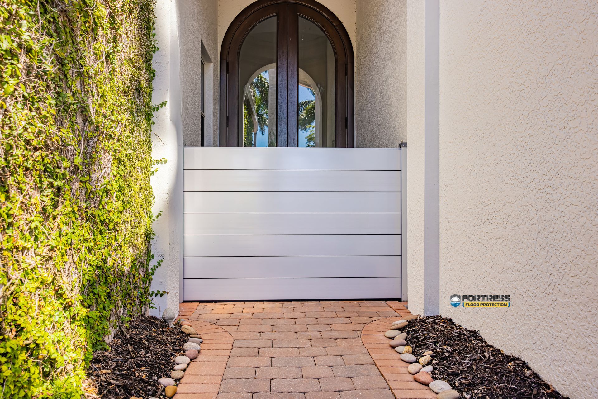 A white flood barrier blocks a brick walkway to arched double doors. Vines grow up the left wall.