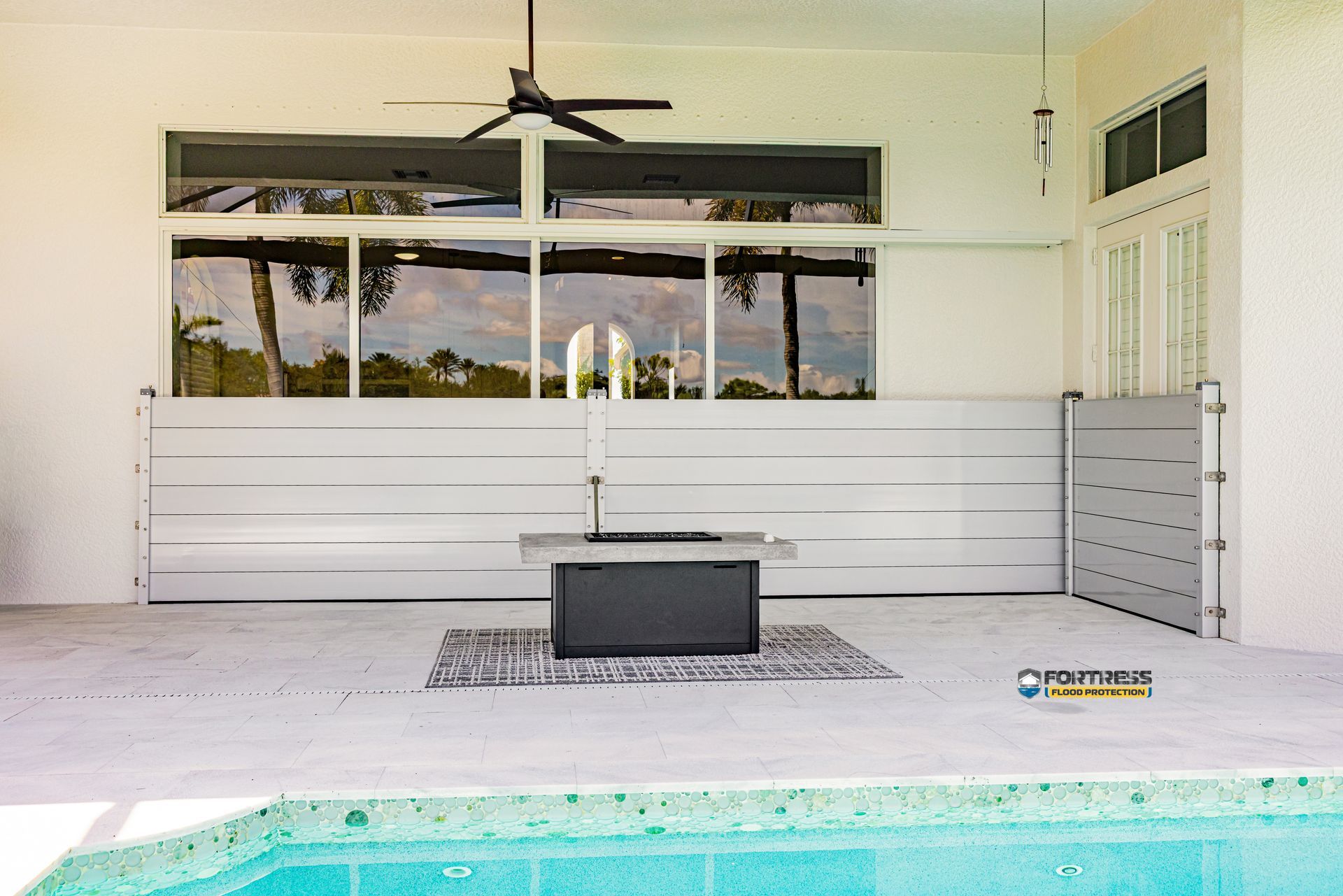White outdoor patio with a horizontal-slat fence, a concrete fire pit, and a rug; palm trees visible through a window.