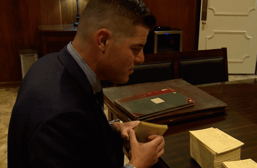 A man in a suit is sitting at a table looking at a book.