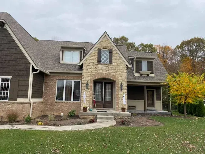 House exterior: stone and brick facade, brown roof, arched entry, fall foliage, cloudy sky.