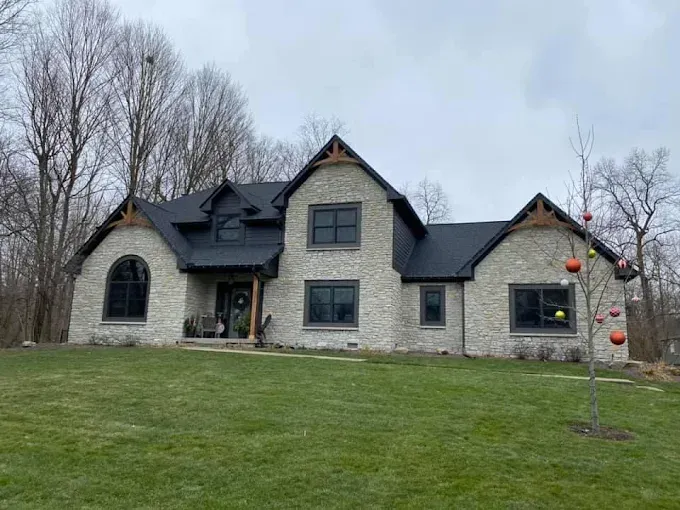 Stone-faced house with black roof and trim, surrounded by a green lawn and trees on a cloudy day.