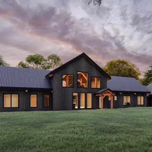 Dark, modern house with black siding, large windows, and a covered entryway, set in a grassy field under a cloudy sky.