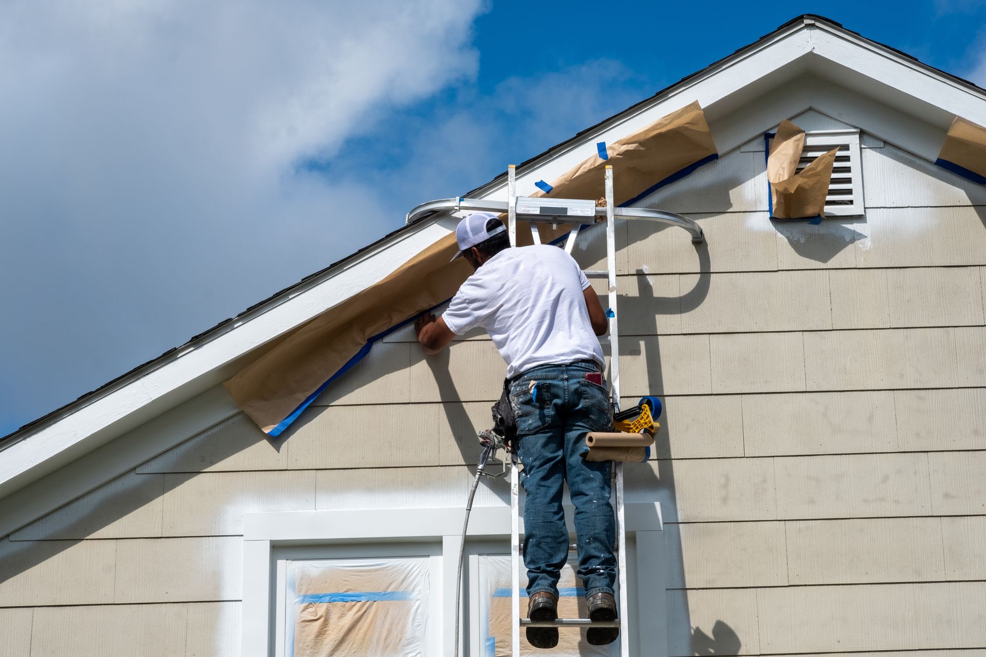 A person on a ladder taping up a house, preparing to paint the trim, blue sky.