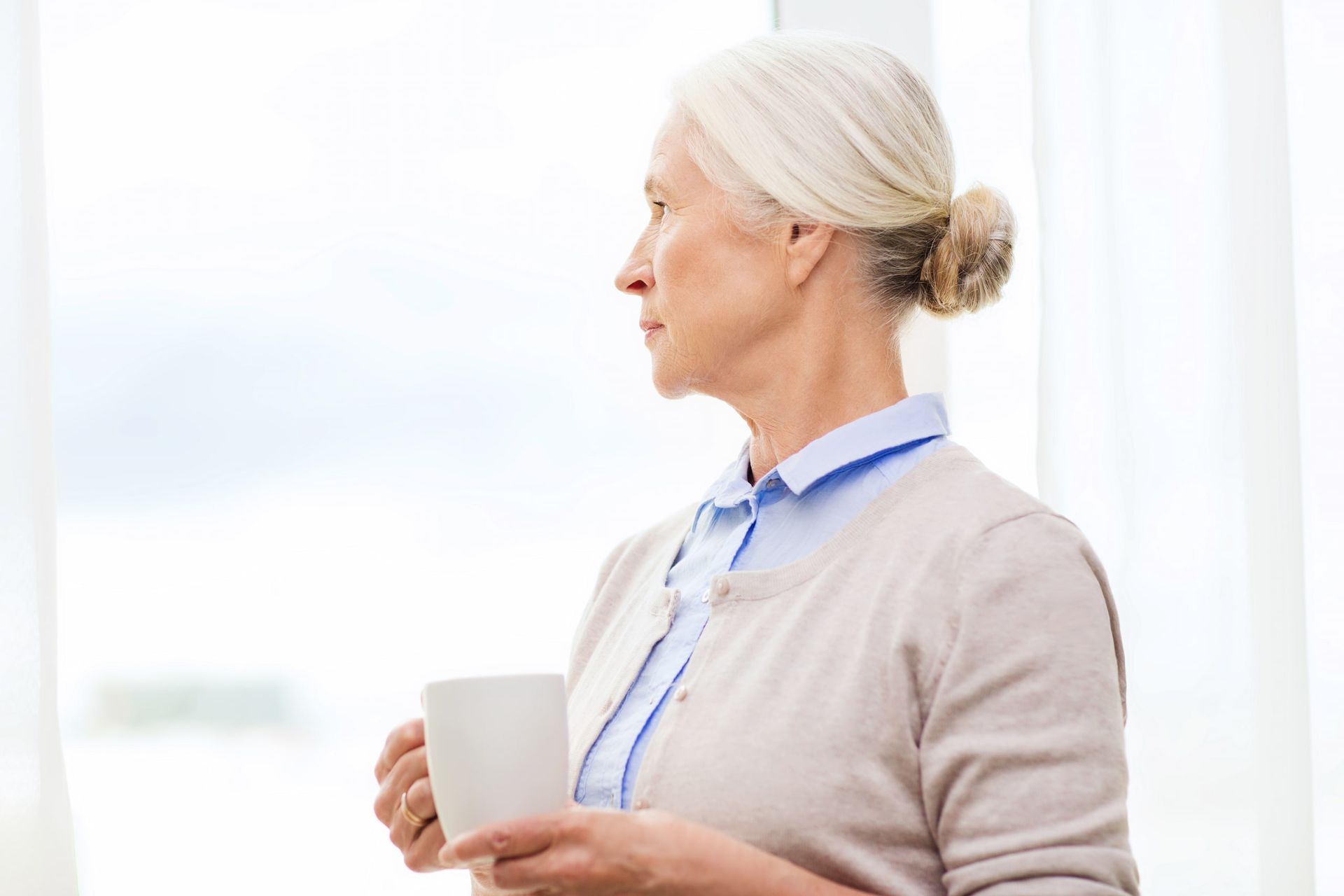 Woman with gray hair, holding a white mug, gazes out a window. She wears a light cardigan and blue shirt.