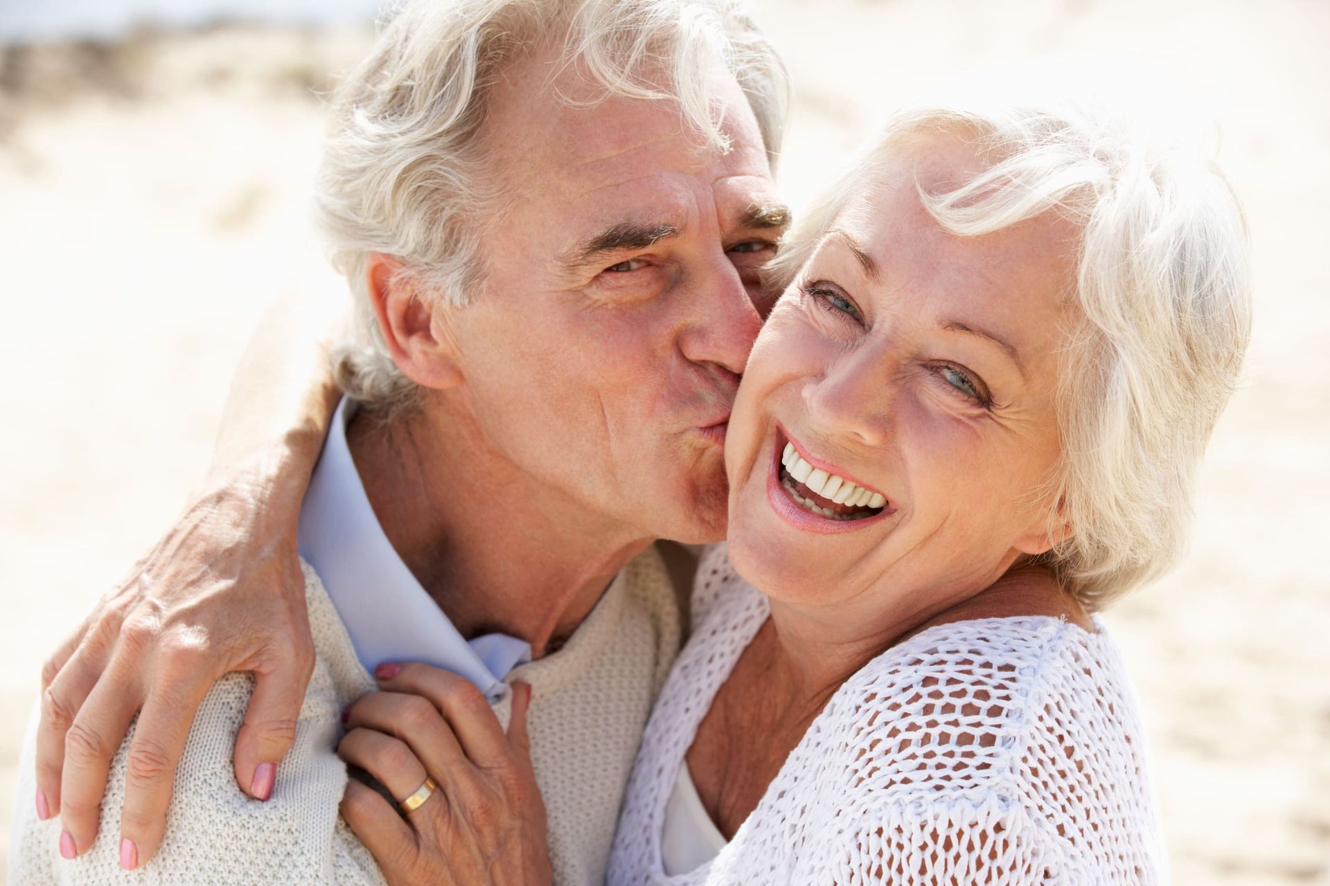 Older man kissing an older woman on the cheek; they are smiling on a beach.