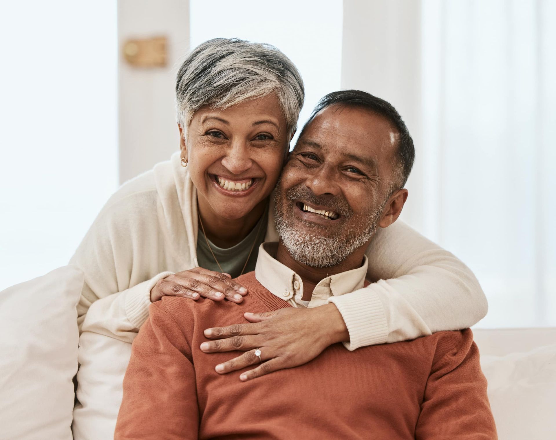 Smiling couple embracing, indoors; woman's arms around man. Warm colors, happy expressions.