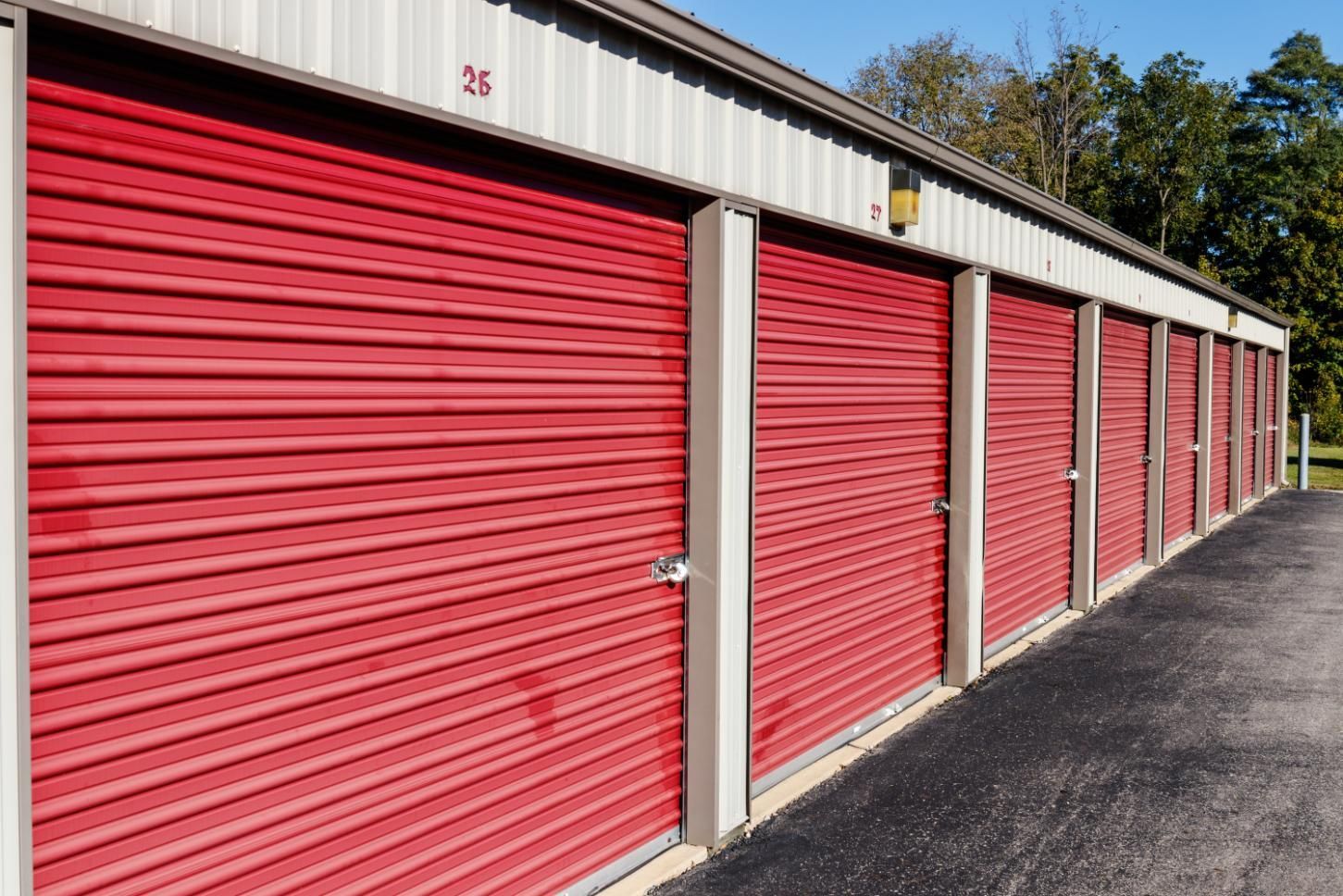 Row of red storage unit doors in a beige building with a black asphalt driveway.