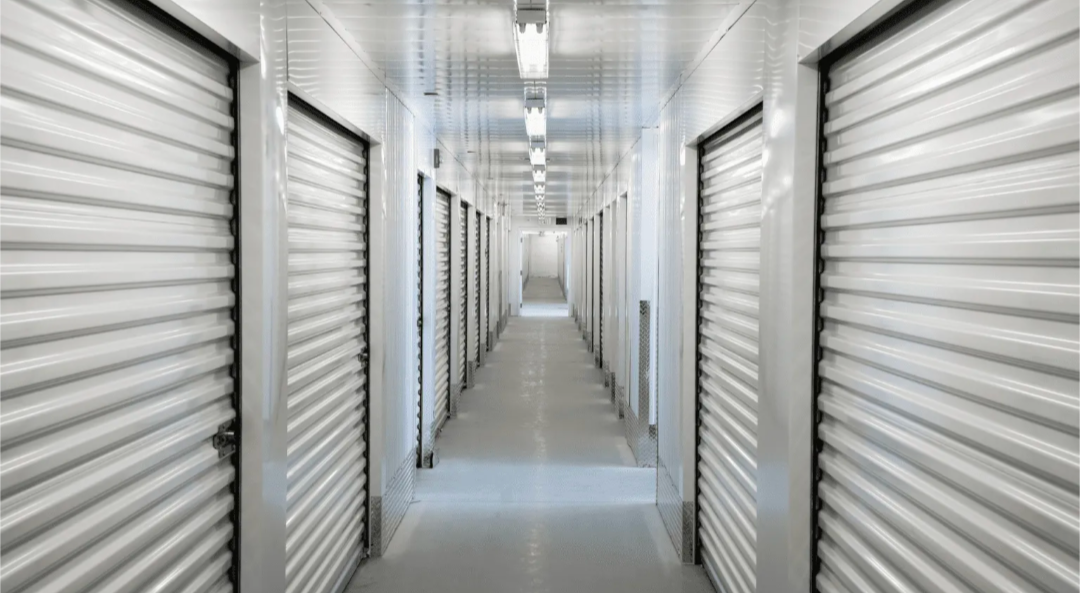 Row of red storage unit doors in a beige building with a black asphalt driveway.