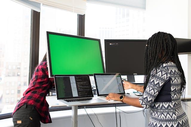 A man and a woman are sitting at a desk with laptops and monitors.