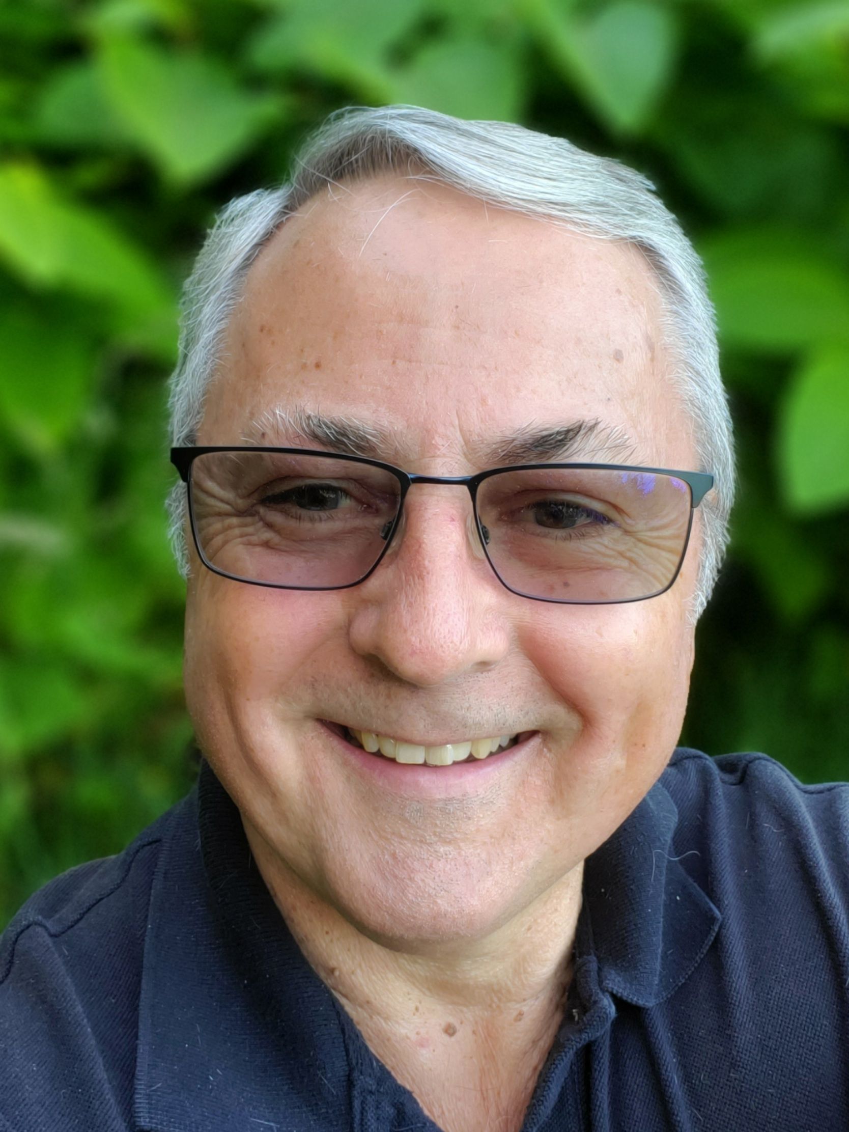 Man with glasses smiling, looking at the camera. He's outdoors with green foliage in the background.