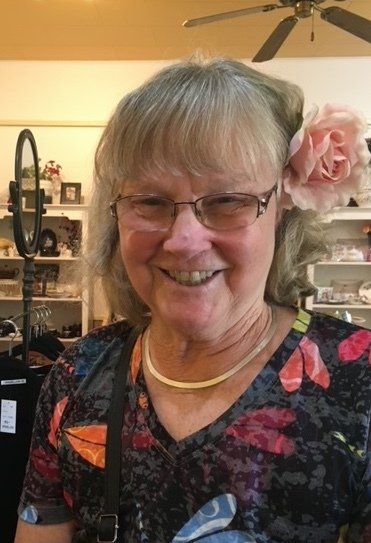 Woman with glasses, pink flower in hair, smiling in a shop with shelves and a fan.