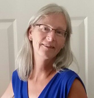 Woman with glasses smiles; wearing a blue sleeveless top, gray hair against white wall.